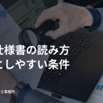 パソコンと資料を虫眼鏡で確認する人の手元。入札仕様書の確認ポイントの見出し入り