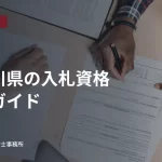 机上の書類を確認し記入する人の手元。神奈川県の入札資格申請ガイドの見出し入り